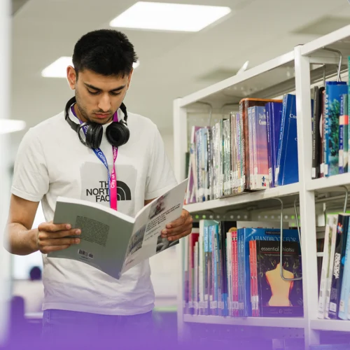 Student is pictured in a library reading the front cover of a book.