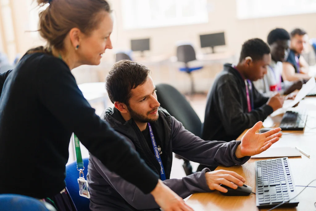 A teacher and a older student discussing something in a classroom setting in front of a computer screen