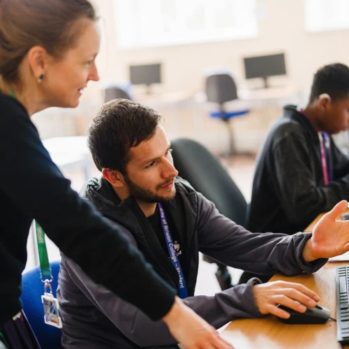A teacher and a older student discussing something in a classroom setting in front of a computer screen
