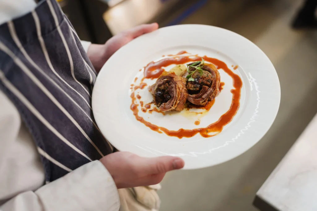 Close-up of a plated bacon-wrapped dish with red sauce and micro herbs, held by a culinary student in a striped apron