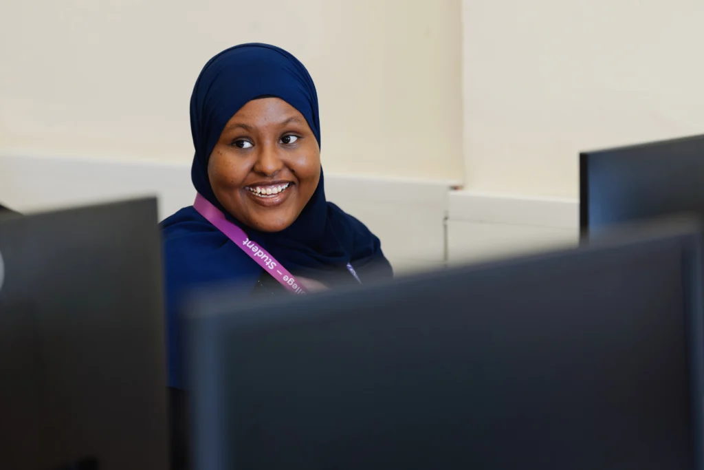 A student is smiling in a classroom setting while on a computer