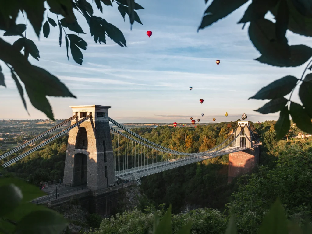 Photo of the historic Clifton Suspension Bridge of Bristol, with hot air balloons flying behind in the distance on a lovely summers day.