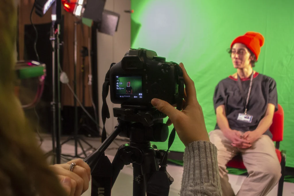 Woman wearing a beanie sitting in front of a camera with green-screen behind her